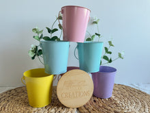 Load image into Gallery viewer, Set of colorful metal buckets with plants on a woven mat, branded 'Little Sun Creations'.
